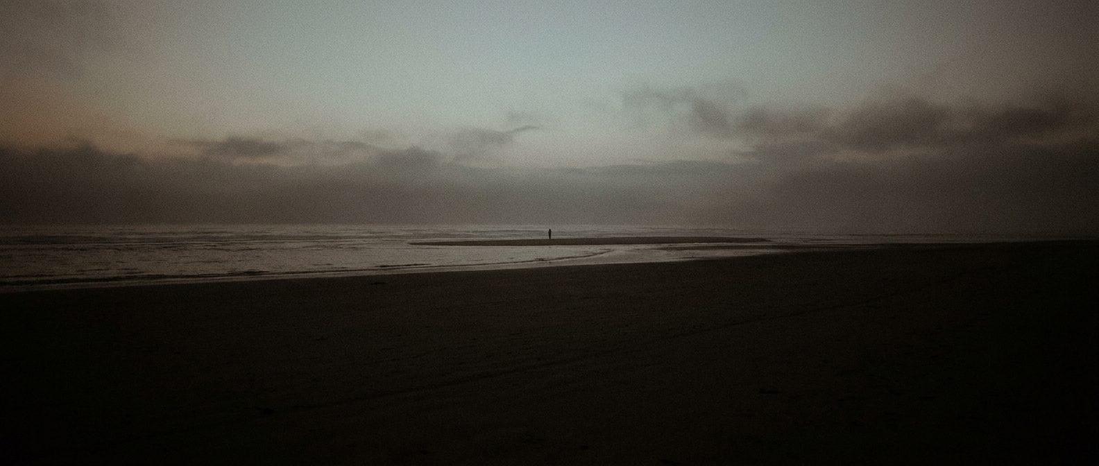 A dark shoreline at dusk, a flat sea meeting a heavy grey sky, near black tonality with a faint warm horizon, vast empty space.